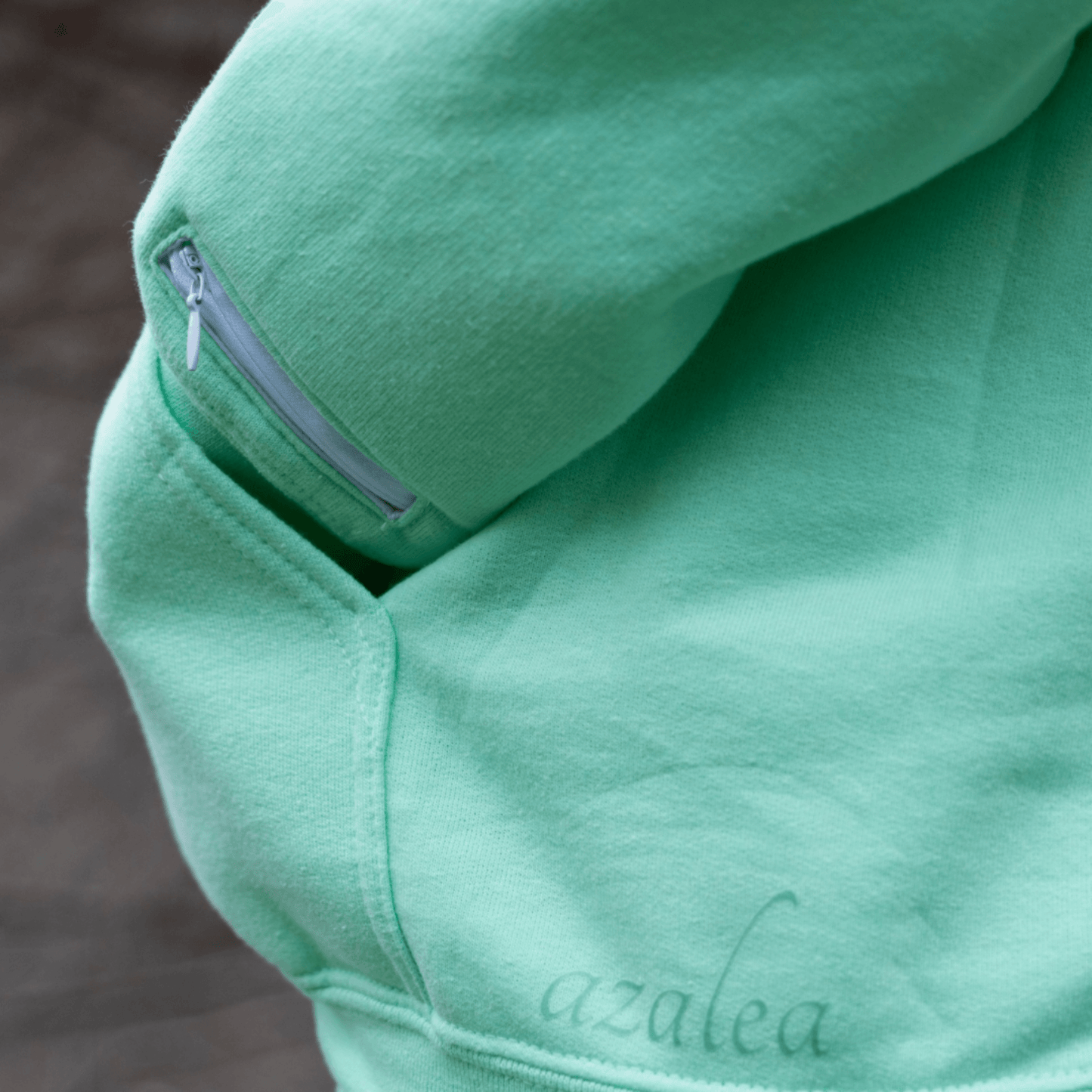 Close-up detail of a mint green Azalea brand comfort hoodie featuring a subtle sleeve pocket with a zipper and the 'azalea' script. This aromatherapy hoodie is designed to gently diffuse calming essential oils, potentially aiding in anxiety relief and promoting mental well-being through its subtle, scented comfort