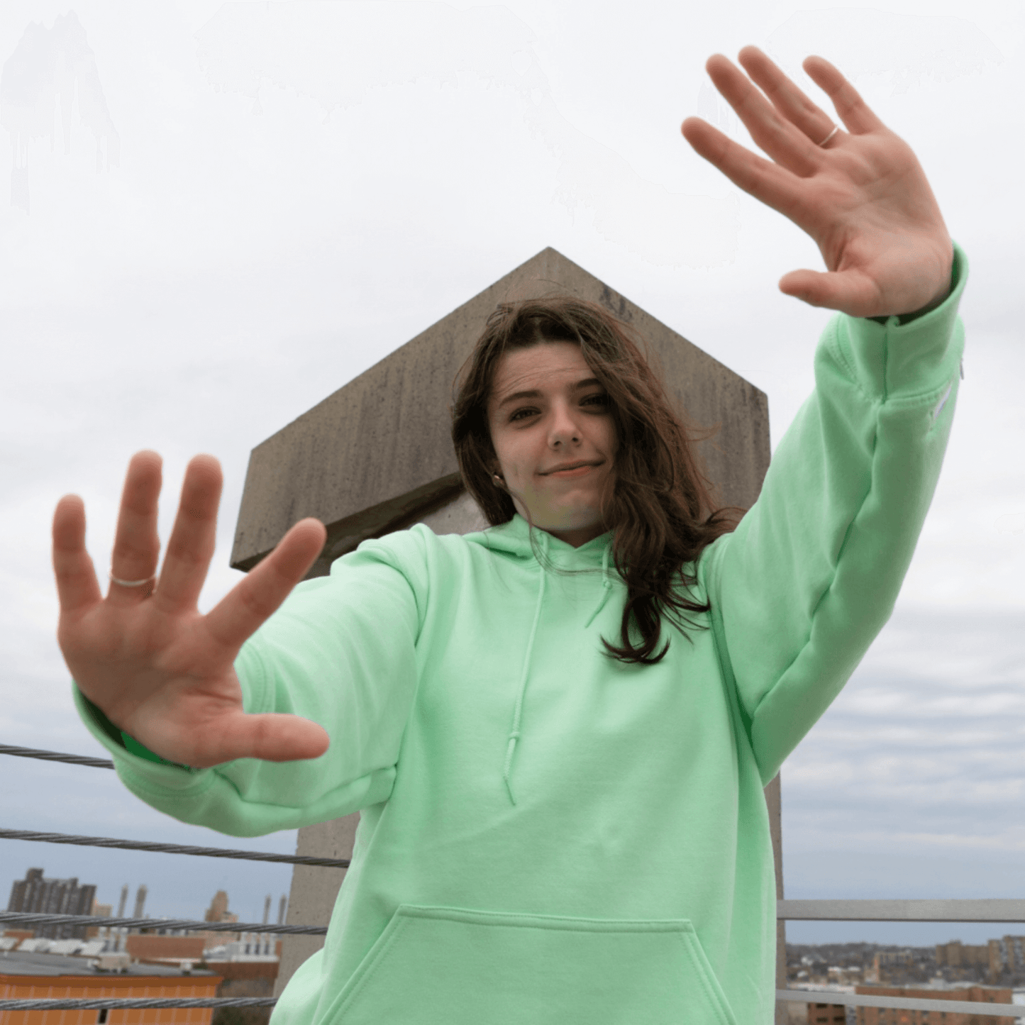 Smiling individual wearing a soft mint green Azalea brand comfort hoodie, captured on an urban rooftop. Infused with natural essential oils, this aromatic apparel offers a fresh, calming scent, creating a unique sensory experience that elevates everyday scenes and promotes a feeling of well-being
